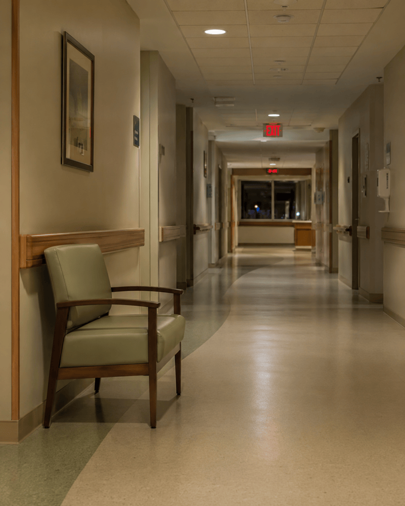 An empty hospital corridor at night with one chair against the wall under fluorescent light, representing medical trauma and the parents who wait there.