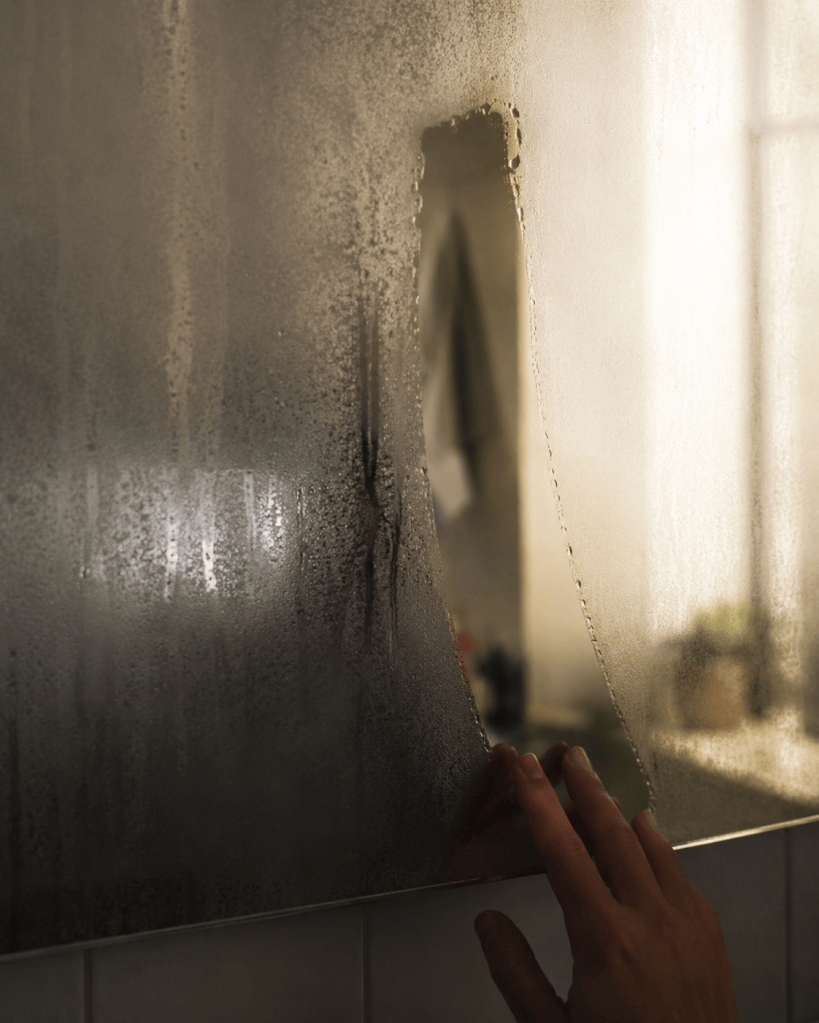  Fogged bathroom mirror with one clear wiped streak showing a blurred partial reflection, soft morning light, muted tones, no identifiable face, quiet editorial still life photography